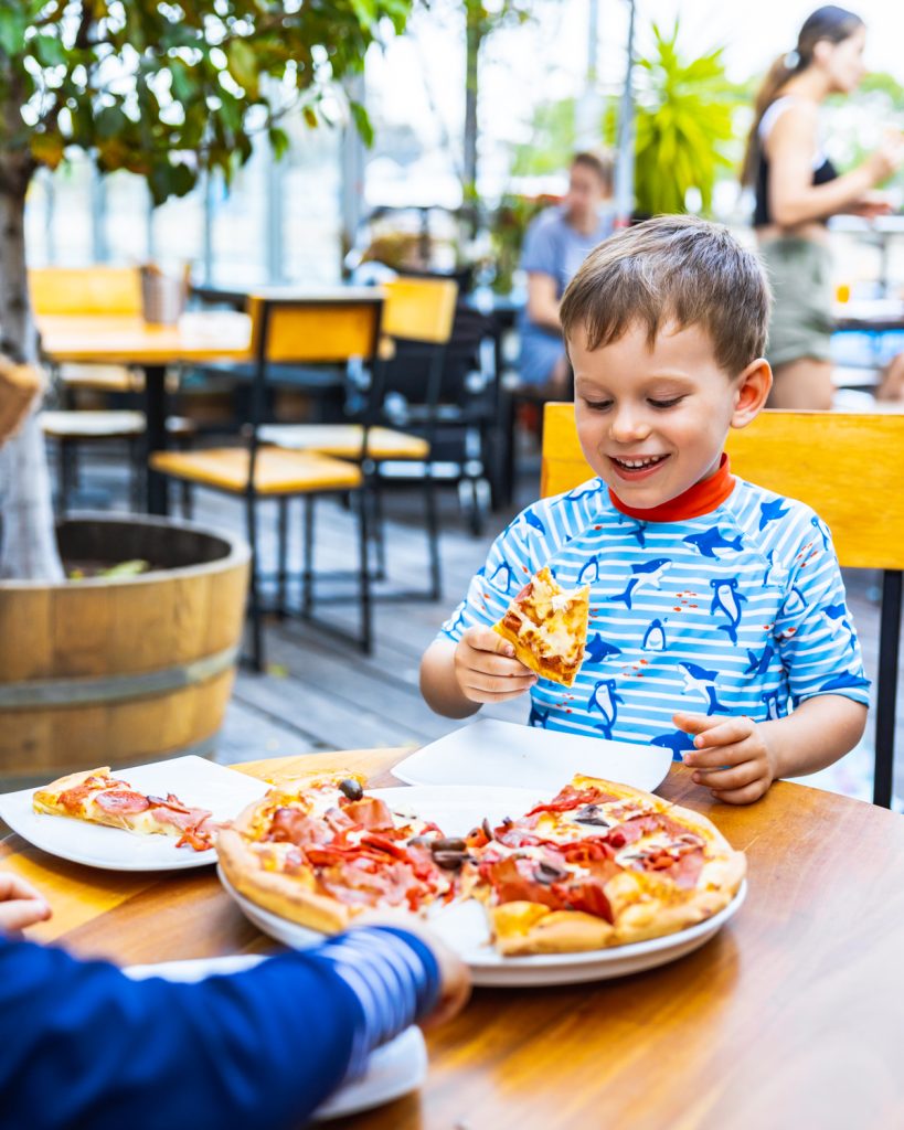 A young boy in a blue shark-patterned rash guard smiles while holding a slice of pizza at an outdoor restaurant.