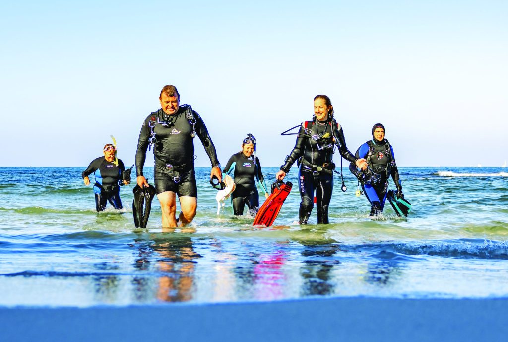 A group of five scuba divers in wetsuits and gear emerge from the ocean, walking towards the shore.