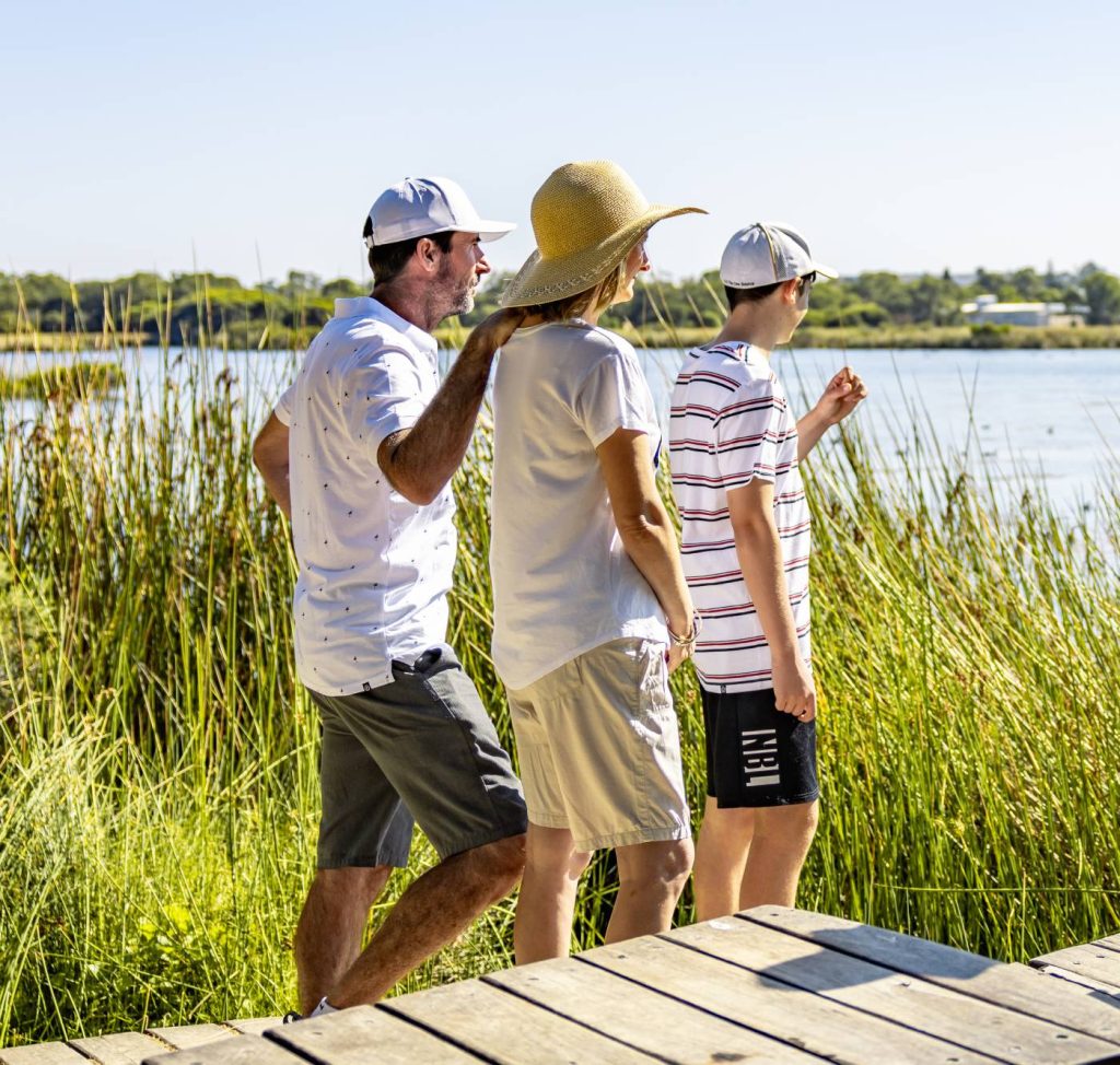 A family of three, wearing summer clothing and hats, walks along a wooden boardwalk near a lake surrounded by tall green reeds.