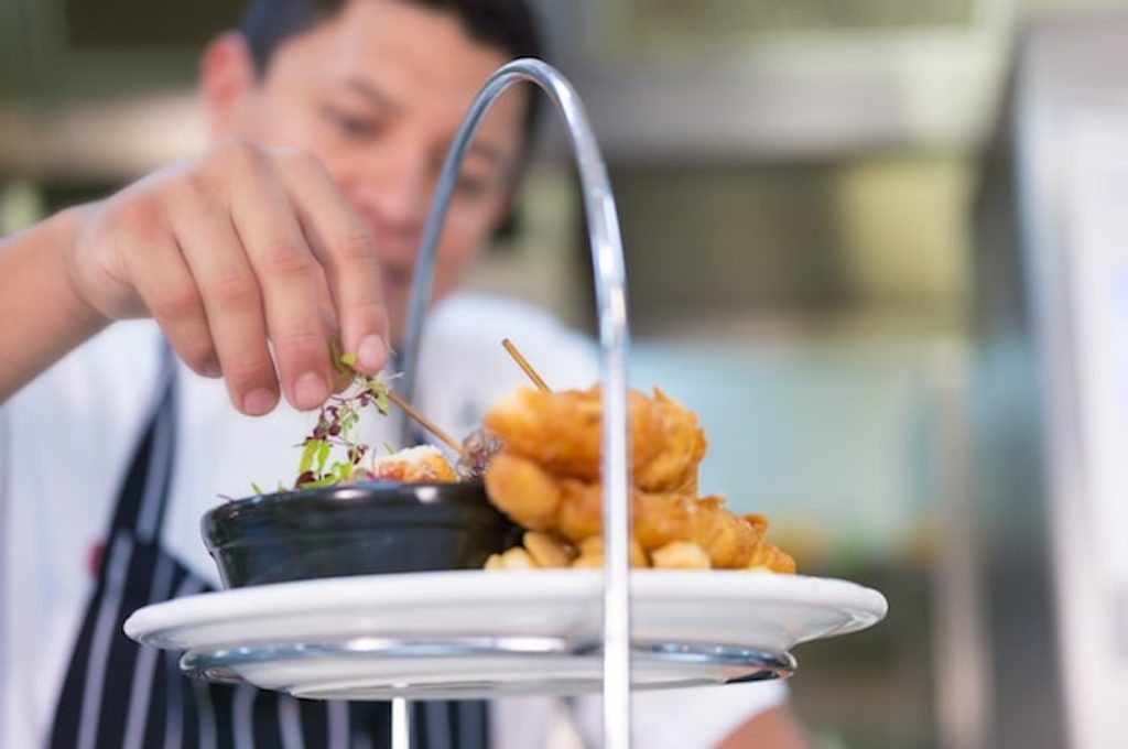 A chef carefully garnishes a dish on a two-tiered serving platter with fried appetizers.