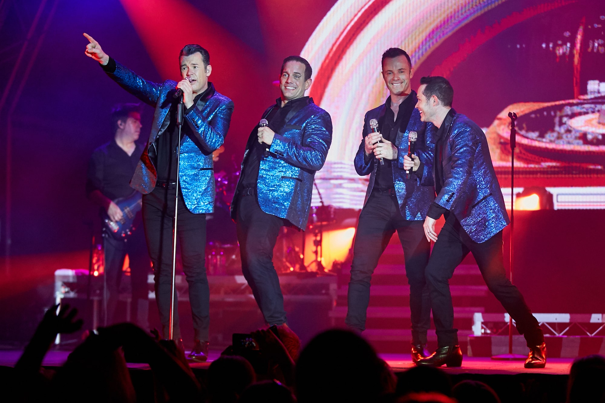 Four male singers in matching blue sequined jackets performing energetically on stage.