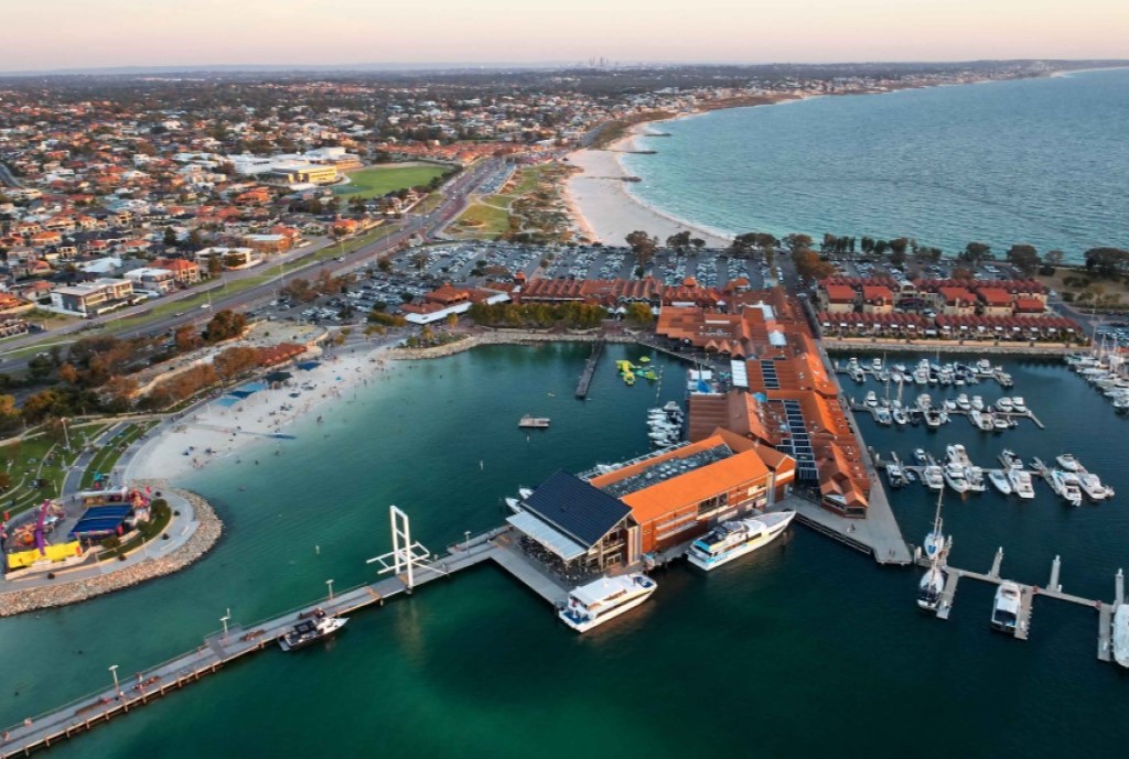 Aerial view of Hillarys Boat Harbour with boats, a boardwalk, a beach, and a surrounding urban landscape at sunset.